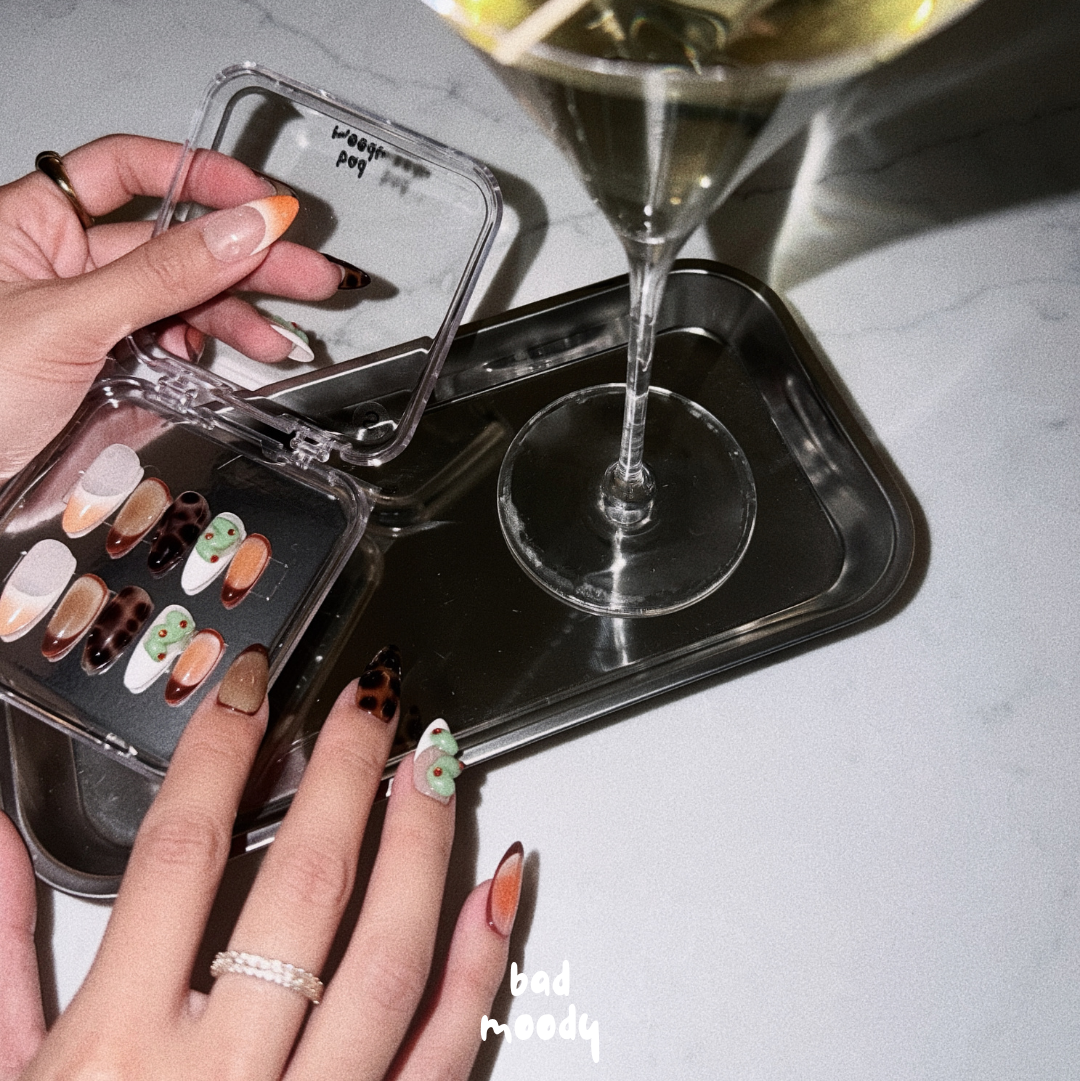 Hand holding a compact mirror with nail polish samples next to a cocktail with olives on a white surface.