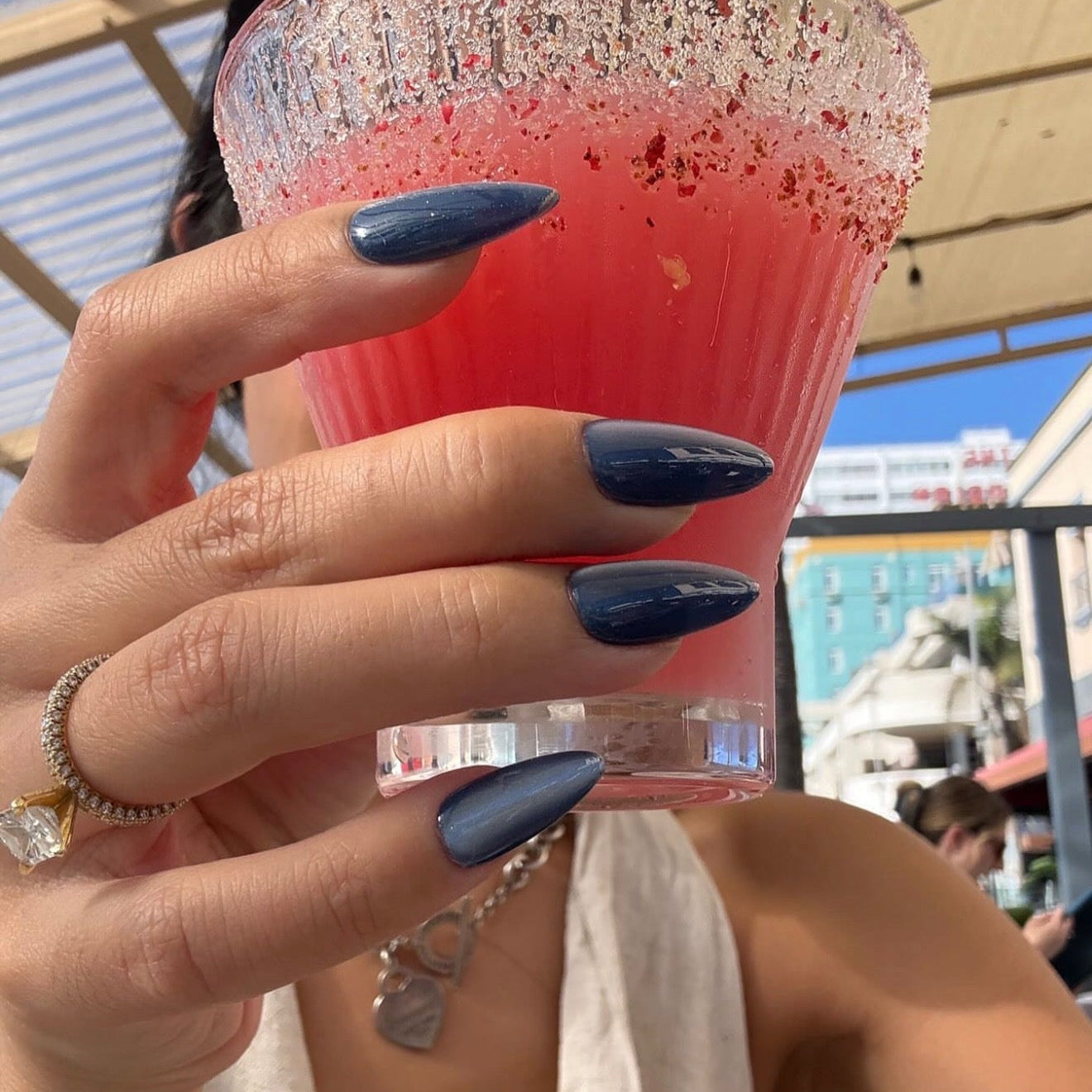 Hand holding a pink drink with a glittery rim, wearing a ring and bracelet, against an outdoor background.